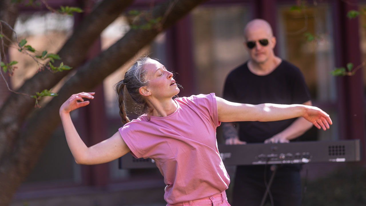 Person dansar utomhus medan en musiker spelar keyboard i bakgrunden.